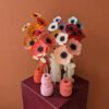 Bouquet d'anémones colorées en papier artisanal disposées dans des vases design sur un socle cubique bordeaux, fond mur terracotta lisse.
