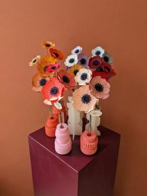 Bouquet d'anémones colorées en papier artisanal disposées dans des vases design sur un socle cubique bordeaux, fond mur terracotta lisse.