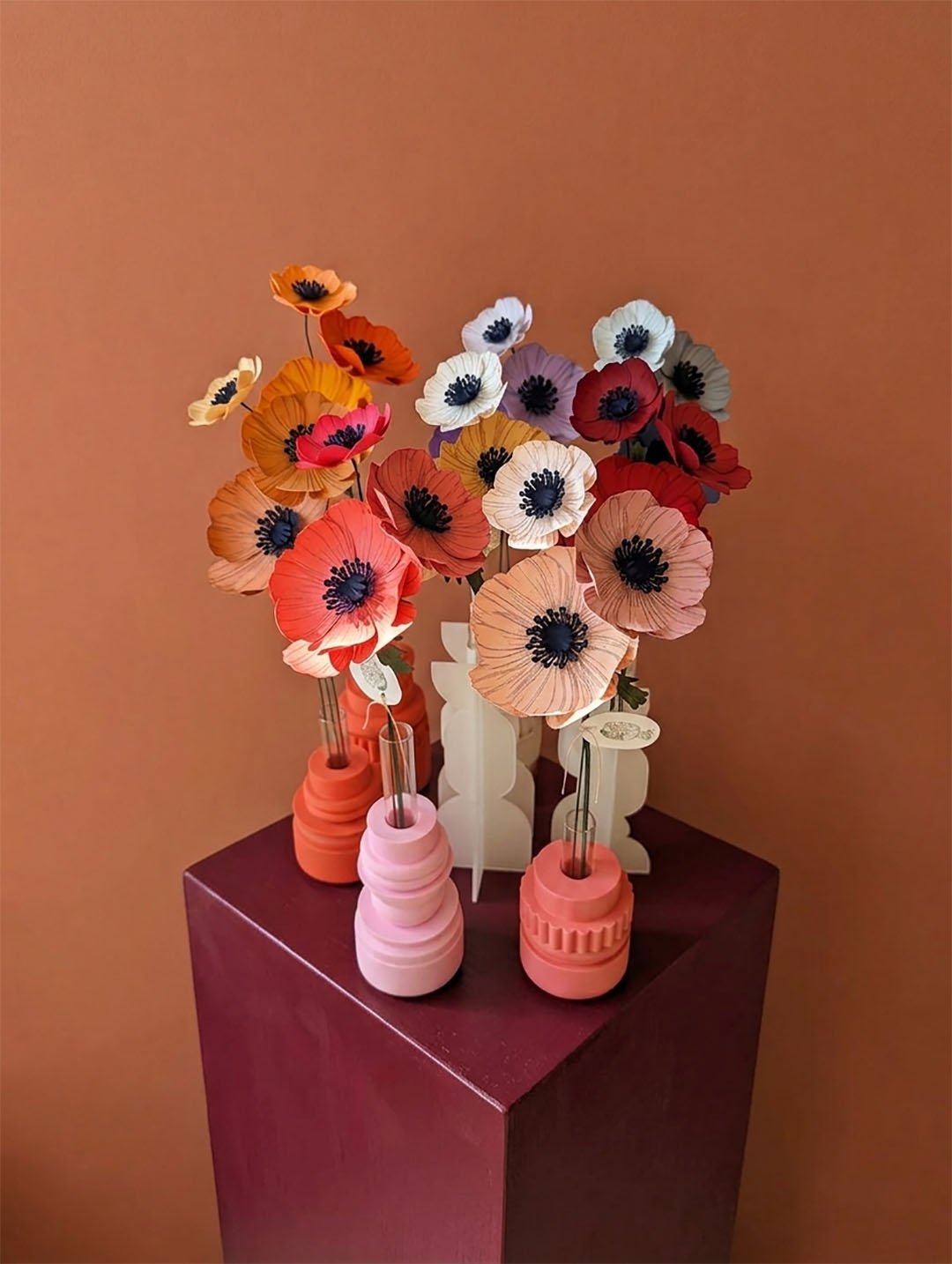 Bouquet d'anémones colorées en papier artisanal disposées dans des vases design sur un socle cubique bordeaux, fond mur terracotta lisse.