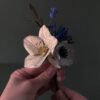 Bouquet de fleurs en papier tenu en main, composé d'une hellébore ivoire, d'une mini anémone blanc et d'un mini chardon bleu outremer