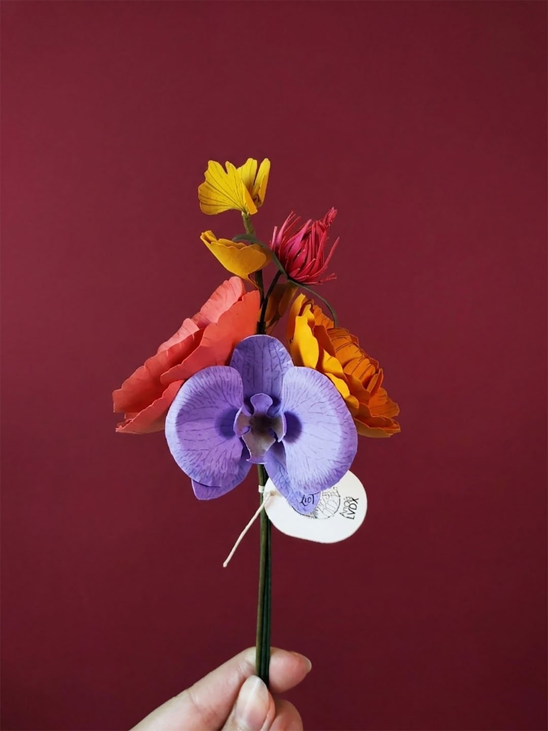 Bouquet de fleurs artisanales en papier d'art coloré, orchidée violette et pavot corail, présenté dans un vase noir sur fond bordeaux. Note : C'est ce qui aide Google Images à comprendre le contenu de la photo et améliore l'accessibilité.