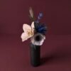 Composition florale artisanale en papier d’art teinté dans la masse, nuances de bleu et beige dans un vase minimaliste, fond vieux rose vénitien sombre.