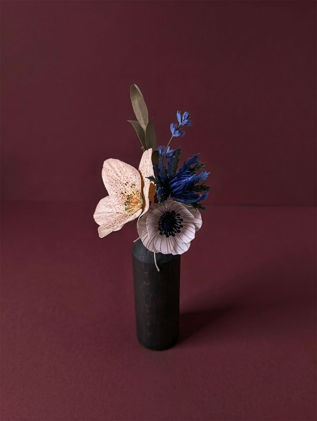 Composition florale artisanale en papier d’art teinté dans la masse, nuances de bleu et beige dans un vase minimaliste, fond vieux rose vénitien sombre.
