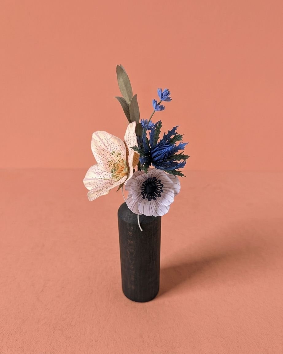 Petit bouquet de fleurs en papier d'art avec une hellébore ivoire, une anémone rose pâle, un chardon bleu, de la mini lavande et du mini olivier, dans un vase en bois sombre