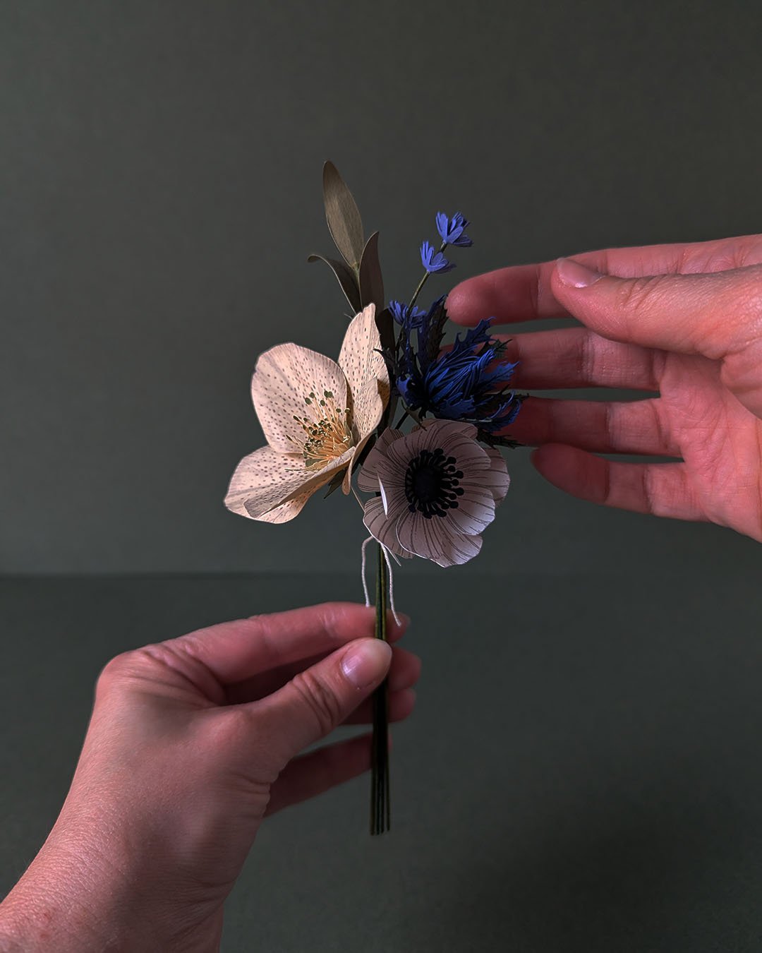 Bouquet de fleurs en papier tenu à deux mains, montrant la composition complète avec hellébore ivoire, anémone blanc et chardon bleu outremer