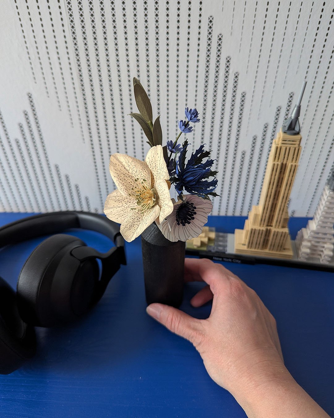 Main tenant un bouquet de fleurs en papier composé d'une hellébore ivoire et d'une anémone blanc, devant une maquette Lego New York sur fond bleu