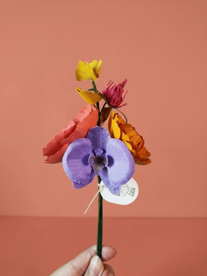 Main tenant un bouquet coloré de fleurs en papier : orchidée iris, renoncule, pavot corail, trèfle viola et ginkgo