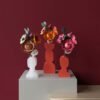 Trois bouquets de fleurs en papier artisanal (renoncule, anémone, capucine) dans des vases géométriques modernes sur un socle épuré, fond uni bordeaux.