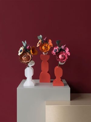 Trois bouquets de fleurs en papier artisanal (renoncule, anémone, capucine) dans des vases géométriques modernes sur un socle épuré, fond uni bordeaux.