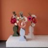 Bouquets de fleurs en papier artisanal dans des vases aux formes géométriques, posés sur une console en terrazzo devant un mur terracotta et un sol en béton ciré anthracite.