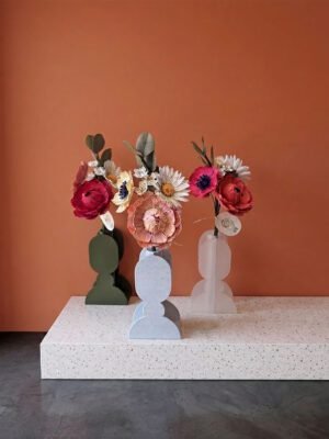 Bouquets de fleurs en papier artisanal dans des vases aux formes géométriques, posés sur une console en terrazzo devant un mur terracotta et un sol en béton ciré anthracite.