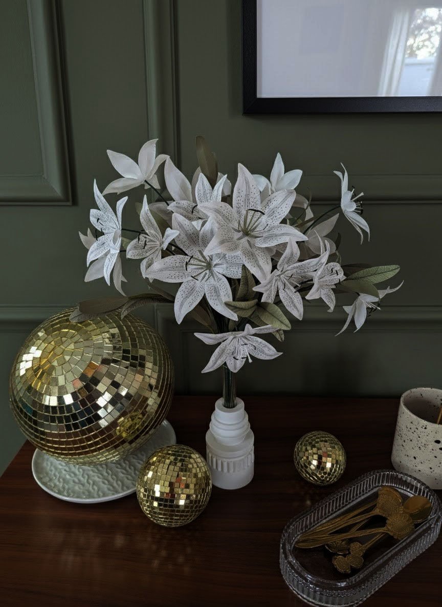 Un bouquet de lys Stargazer en papier fait main, présenté dans un petit vase blanc cannelé sur une table en bois, entouré de boules à facettes dorées.