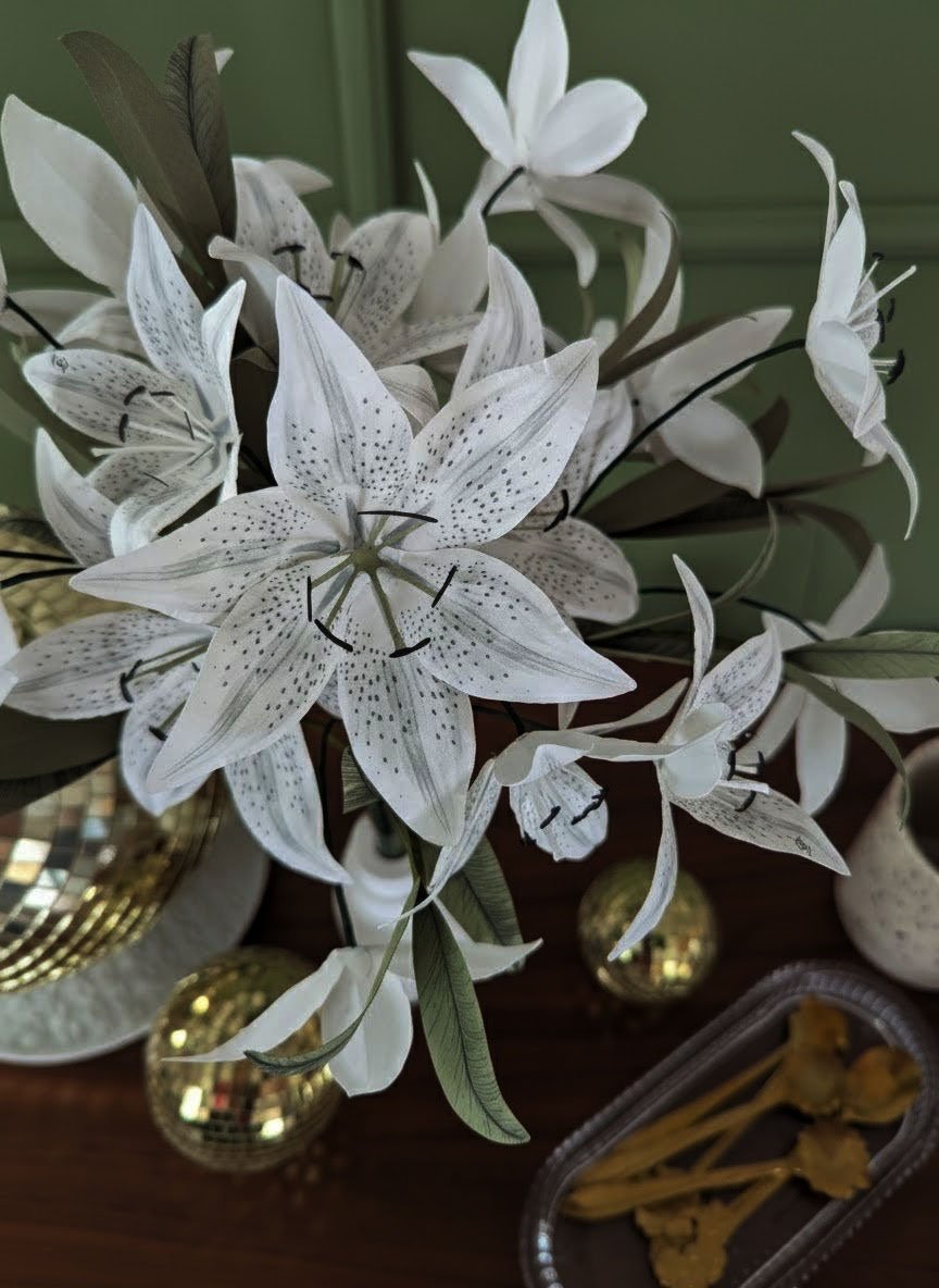 Gros plan sur un lys Stargazer artisanal en papier, montrant les mouchetures grises détaillées et le pistil vert sur un fond de décoration dorée.