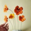 Cinq fleurs de capucine en papier d'art orange et jaune soleil, montées sur tiges, présentées sur un socle en marbre blanc et gris, tenues par une main