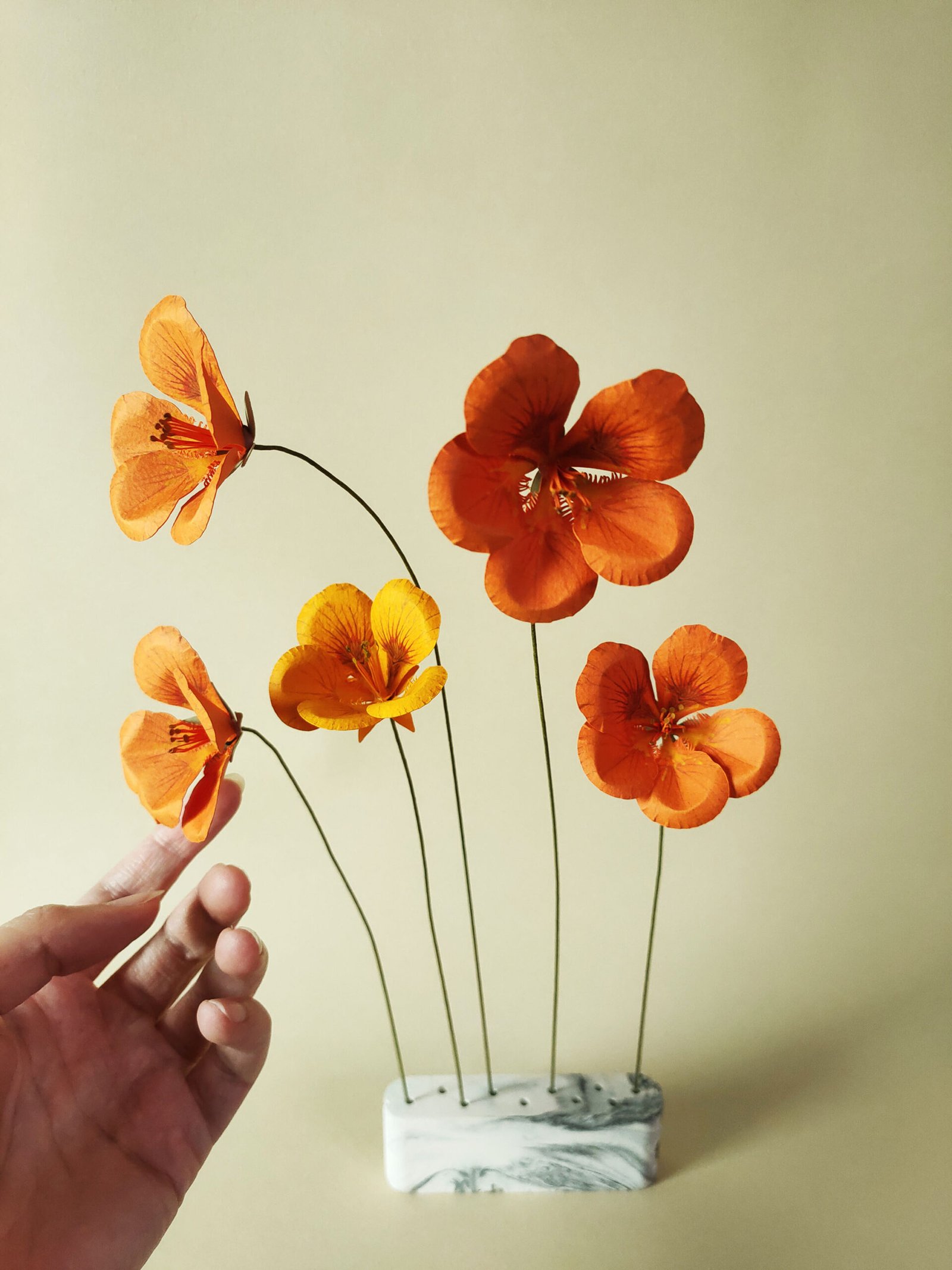 Cinq fleurs de capucine en papier d'art orange et jaune soleil, montées sur tiges, présentées sur un socle en marbre blanc et gris, tenues par une main