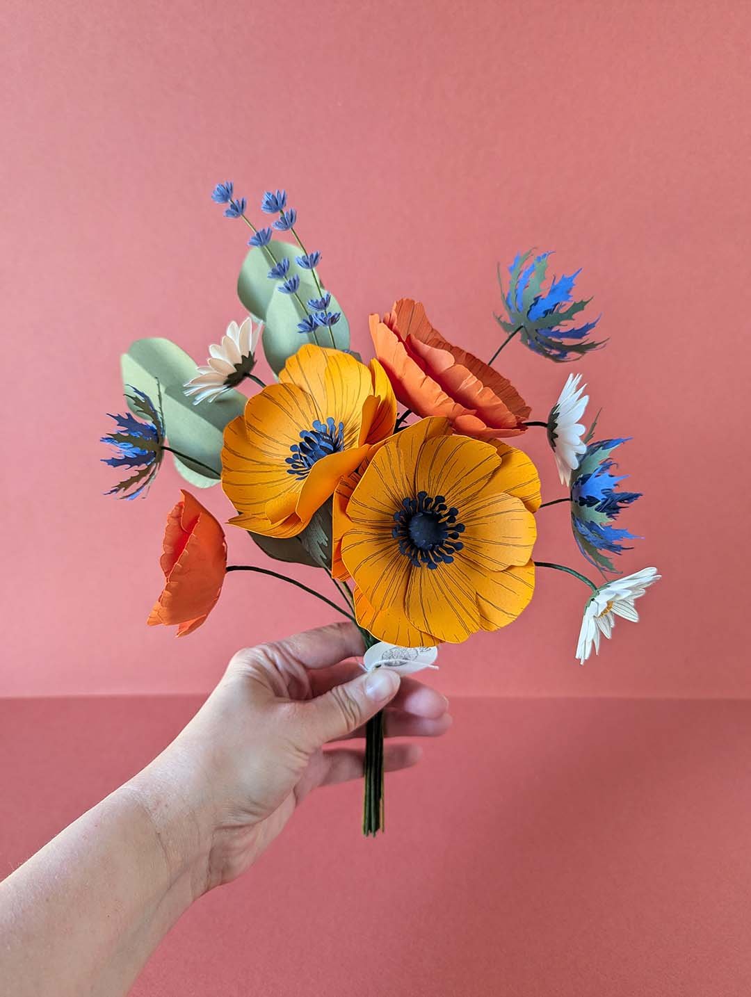 Main tenant le bouquet de fleurs en papier modèle Garrigue, avec anémones jaune soleil, pavots orange, pâquerettes blanches, chardons bleu outremer, lavande et eucalyptus, fond rose