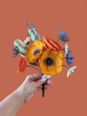 Main tenant le bouquet de fleurs en papier modèle Garrigue, avec anémones jaune soleil, pavots orange, pâquerettes blanches, chardons bleu outremer, lavande et eucalyptus, fond terracotta