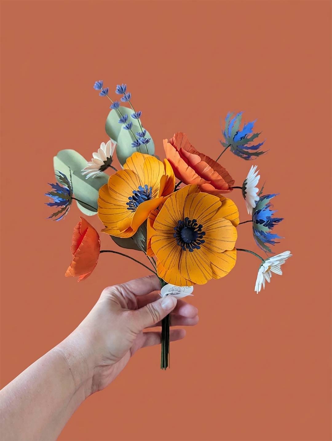 Main tenant le bouquet de fleurs en papier modèle Garrigue, avec anémones jaune soleil, pavots orange, pâquerettes blanches, chardons bleu outremer, lavande et eucalyptus, fond terracotta