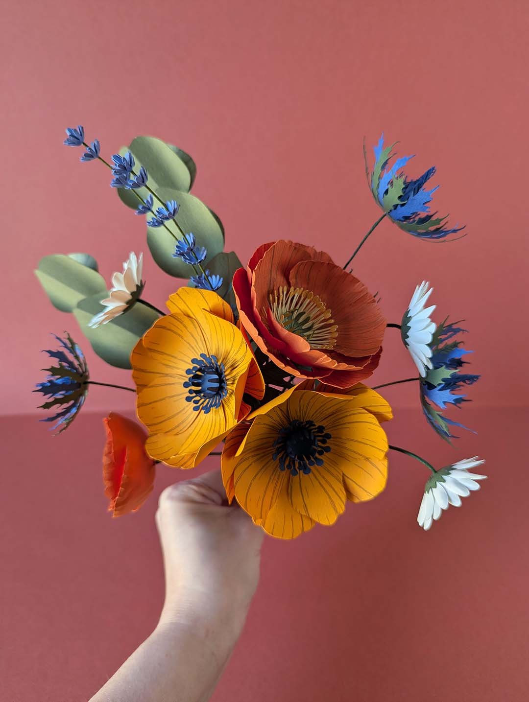 Vue de dessus du bouquet Garrigue en fleurs de papier tenu en main, avec anémones jaune soleil, pavot rouge-orangé face ouverte, chardons bleu outremer, pâquerettes blanches, lavande et eucalyptus, fond rose