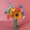 Bouquet de fleurs en papier artisanales modèle Garrigue posé dans un vase vert géométrique, composé de deux grandes anémones jaune soleil, d'un pavot et d'un mini pavot orange, de pâquerettes blanches, de chardons bleu outremer, de brins de lavande et de tiges d'eucalyptus, fond rose