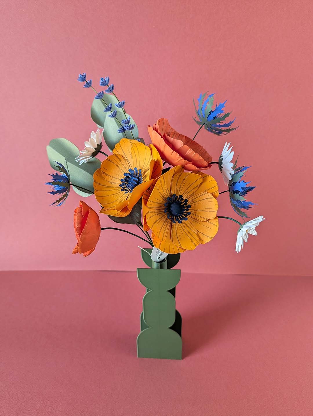 Bouquet de fleurs en papier artisanales modèle Garrigue posé dans un vase vert géométrique, composé de deux grandes anémones jaune soleil, d'un pavot et d'un mini pavot orange, de pâquerettes blanches, de chardons bleu outremer, de brins de lavande et de tiges d'eucalyptus, fond rose