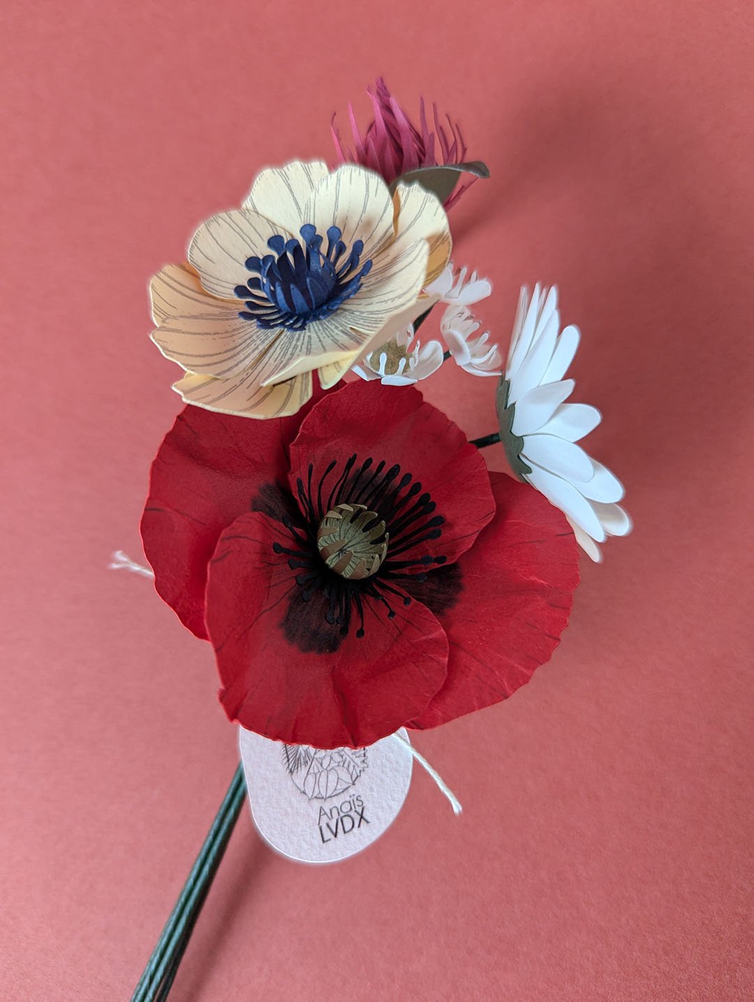 Bouquet de fleurs en papier artisanal tenu debout de biais, coquelicot rouge et anémone crème au premier plan, fond terracotta