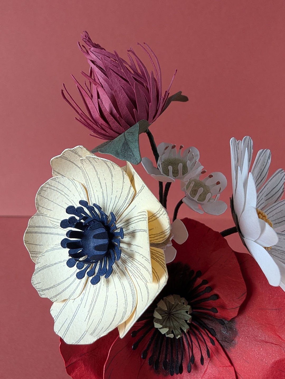 Bouquet de fleurs en papier artisanal avec coquelicot rouge, anémone crème à cœur bleu marine et trèfle bordeaux, fond rose terracotta