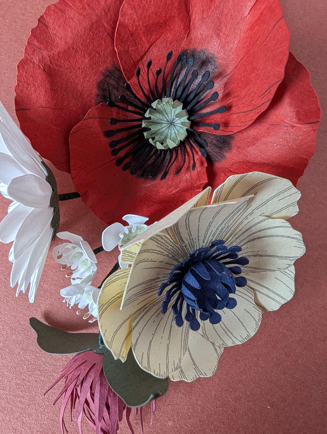 Vue plongeante sur un bouquet de fleurs en papier avec un petit coquelicot rouge et une anémone crème, sur fond terracotta