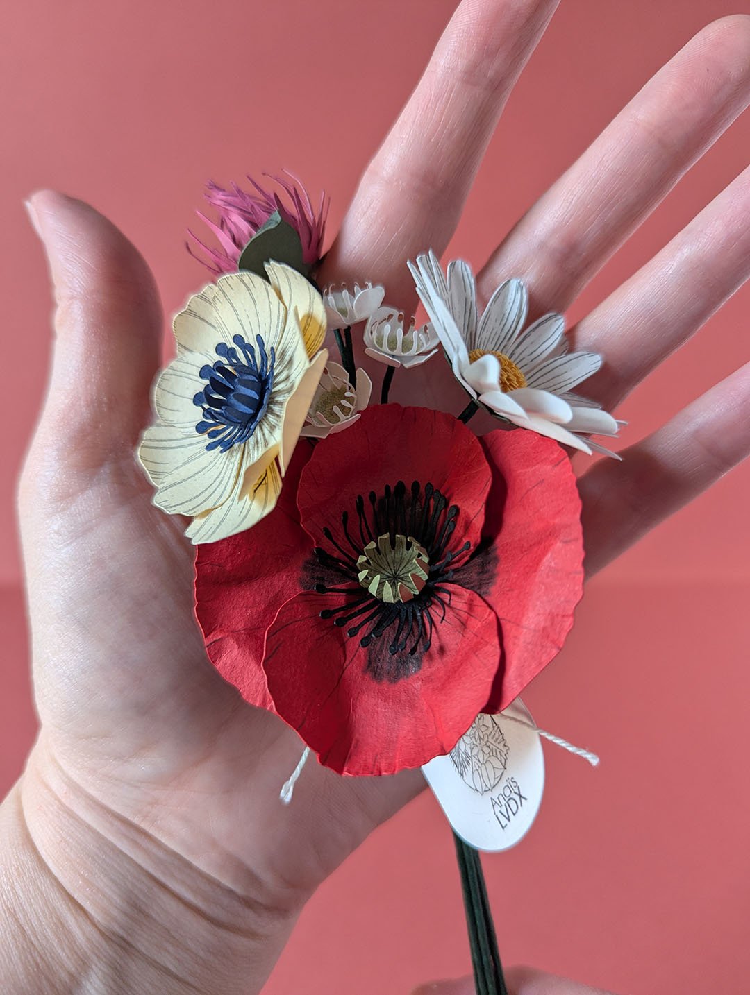 Petit bouquet de fleurs en papier artisanal posé dans une paume ouverte pour montrer sa taille, fond terracotta