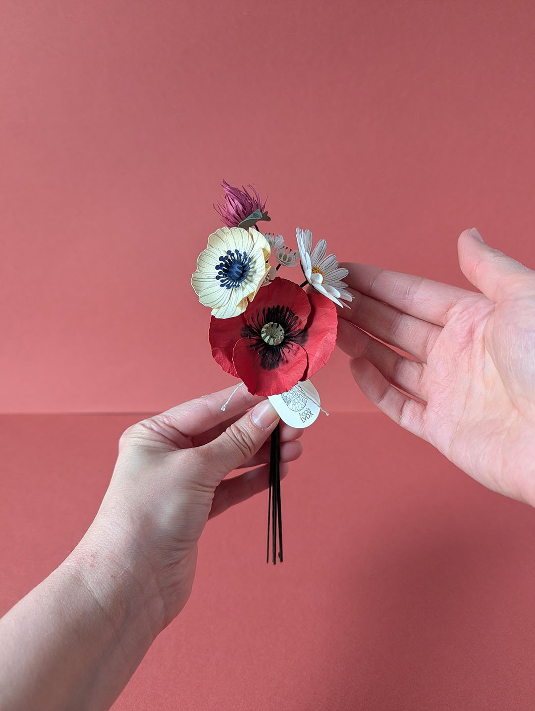 Bouquet de fleurs en papier artisanal présenté tendu entre deux paires de mains, geste d'offrande, fond terracotta