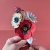 Bouquet de fleurs en papier artisanal tenu à deux mains, coquelicot rouge et anémone crème visibles, fond terracotta