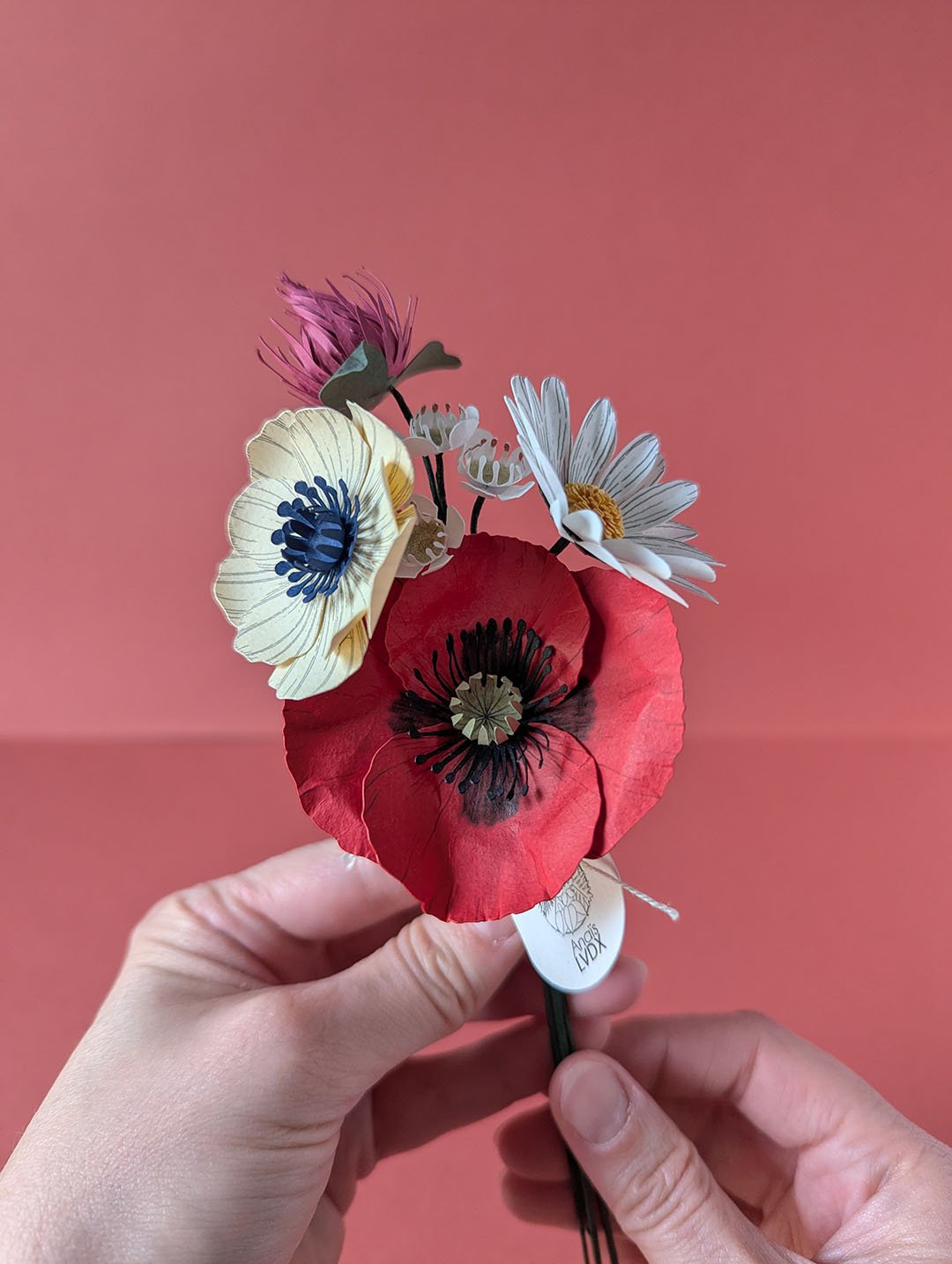 Bouquet de fleurs en papier artisanal tenu à deux mains, coquelicot rouge et anémone crème visibles, fond terracotta