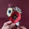 Bouquet de fleurs en papier artisanal tenu à deux mains, coquelicot rouge et anémone crème visibles, fond boredaux