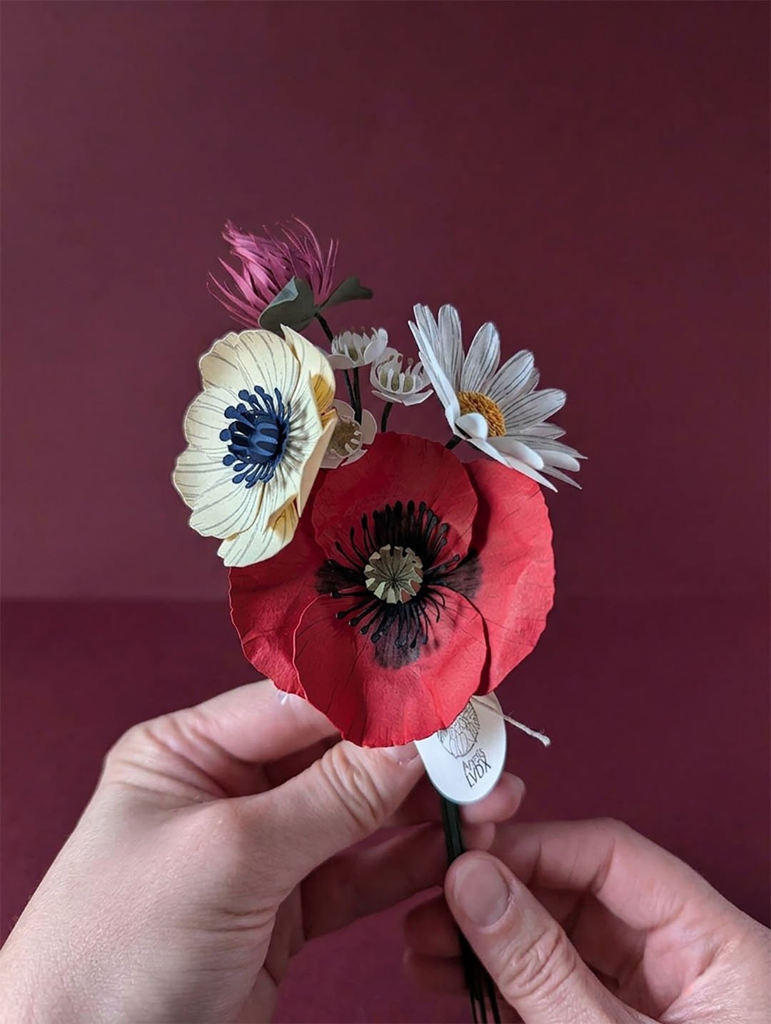 Bouquet de fleurs en papier artisanal tenu à deux mains, coquelicot rouge et anémone crème visibles, fond boredaux