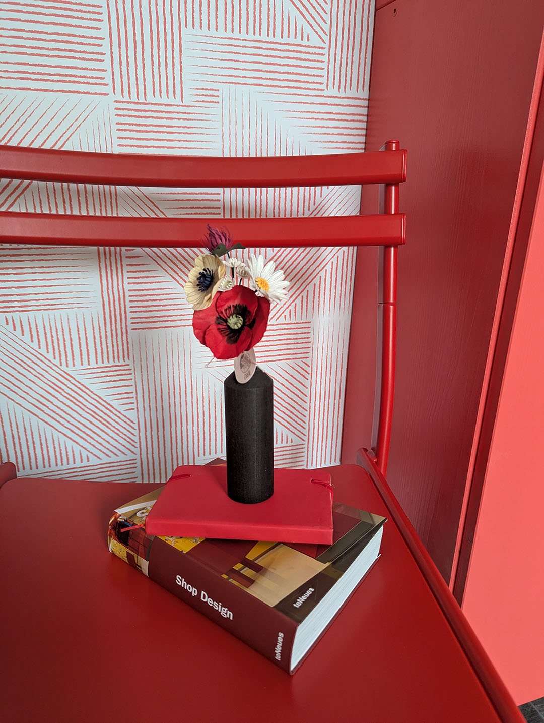 Bouquet de fleurs en papier dans un vase noir cylindrique, posé sur des livres et une chaise rouge, fond papier peint graphique rouge et blanc