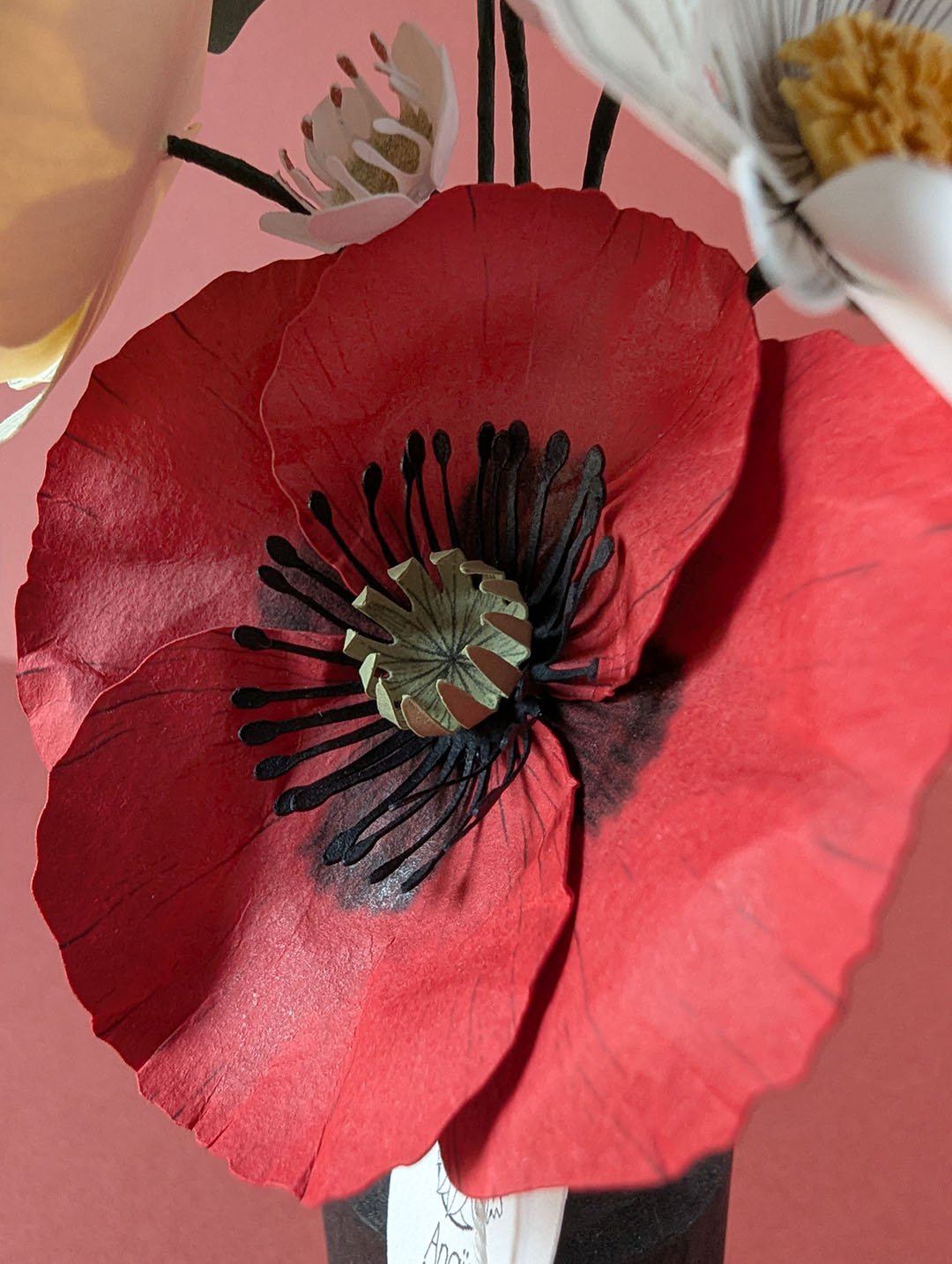 Gros plan sur un coquelicot en papier rouge vif à pistil noir et cœur vert sauge, fond rose poudré