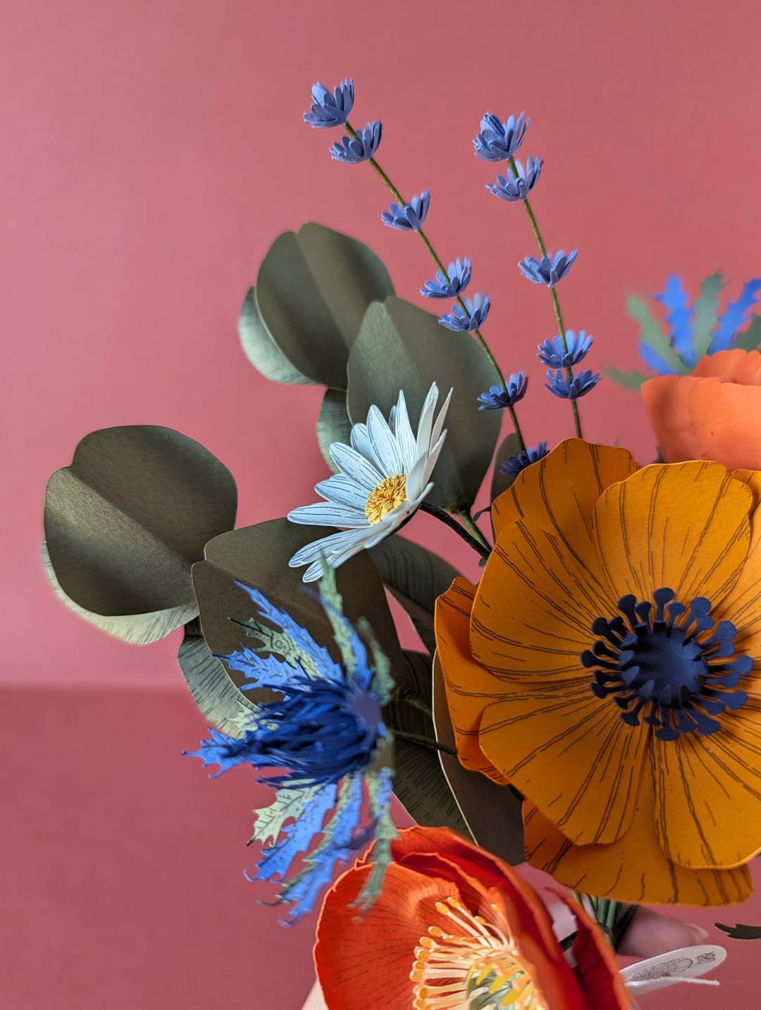 Gros plan sur les brins de lavande bleue, une pâquerette blanche et un chardon bleu outremer du bouquet Garrigue en fleurs de papier, avec feuillage d'eucalyptus vert olive en arrière-plan