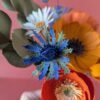 Gros plan sur un chardon bleu outremer aux bractées dentelées vertes du bouquet Garrigue en fleurs de papier, avec un mini pavot orange et une pâquerette blanche en arrière-plan