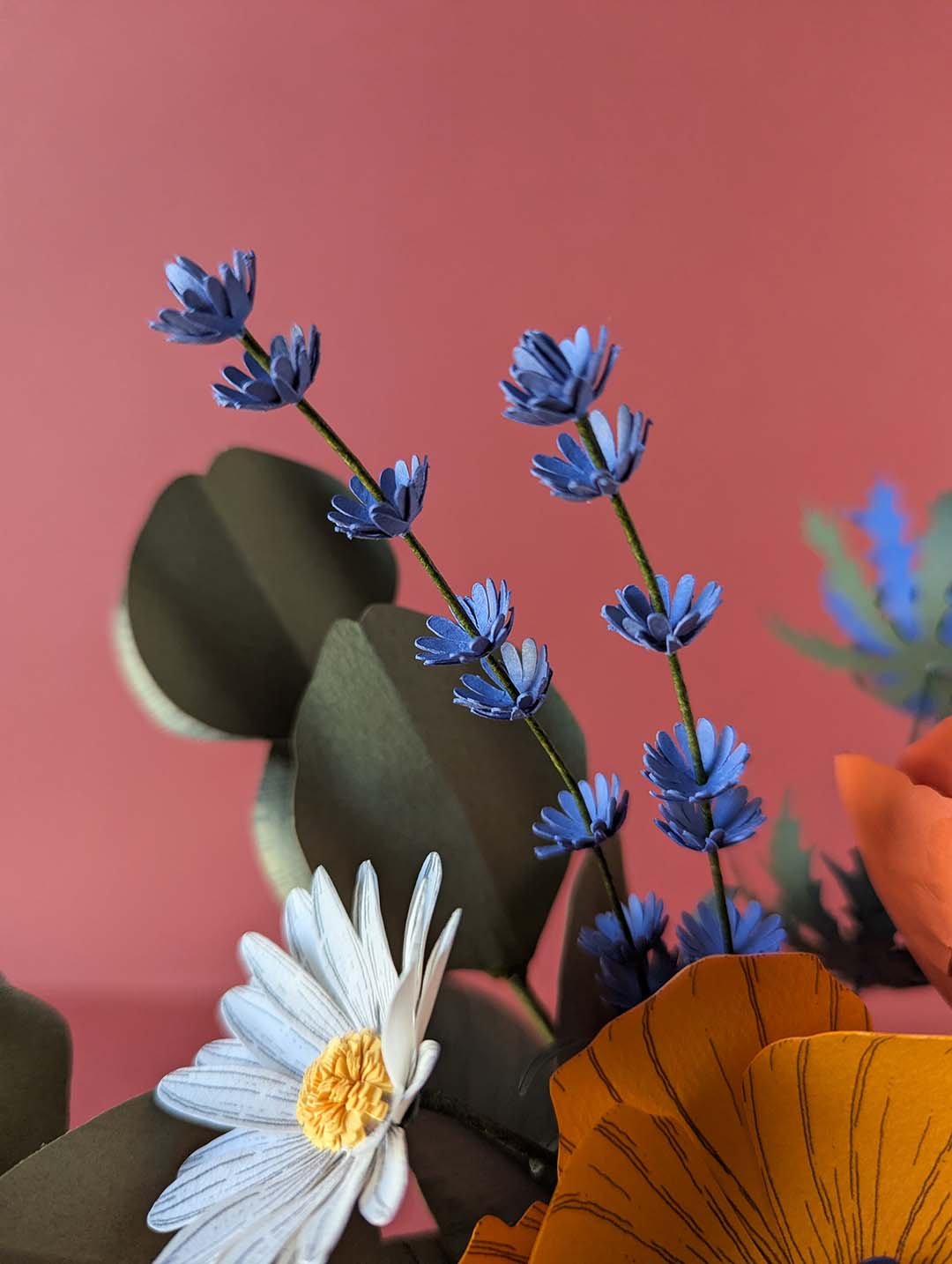 Gros plan sur deux tiges de lavande bleu violet et une pâquerette blanche au cœur jaune du bouquet Garrigue en fleurs de papier artisanales, fond rose