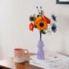 Bouquet Garrigue en papier d'art teinté dans la masse, composé d'anémones jaune soleil, pavots orange, chardons bleus et lavande, dans un vase lilas géométrique sur commode en bois, Anaïs LVDX