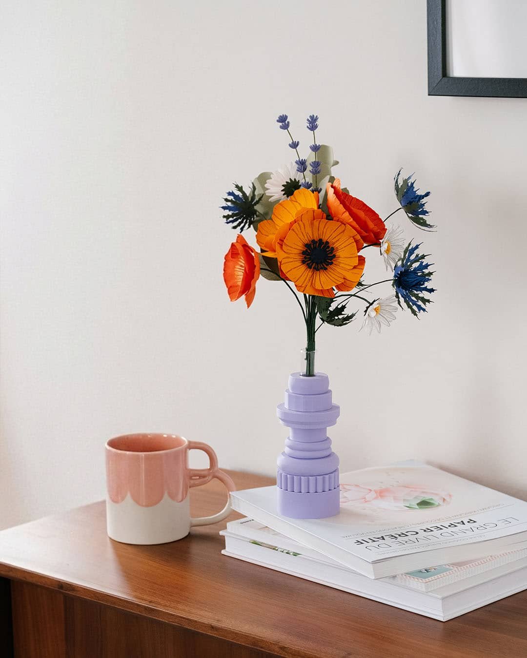 Bouquet Garrigue en papier d'art teinté dans la masse, composé d'anémones jaune soleil, pavots orange, chardons bleus et lavande, dans un vase lilas géométrique sur commode en bois, Anaïs LVDX