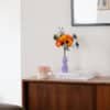 Bouquet de fleurs en papier d'art Garrigue dans un vase lilas posé sur des livres, sur une commode en bois foncé, poster botanique encadré en arrière-plan, Anaïs LVDX