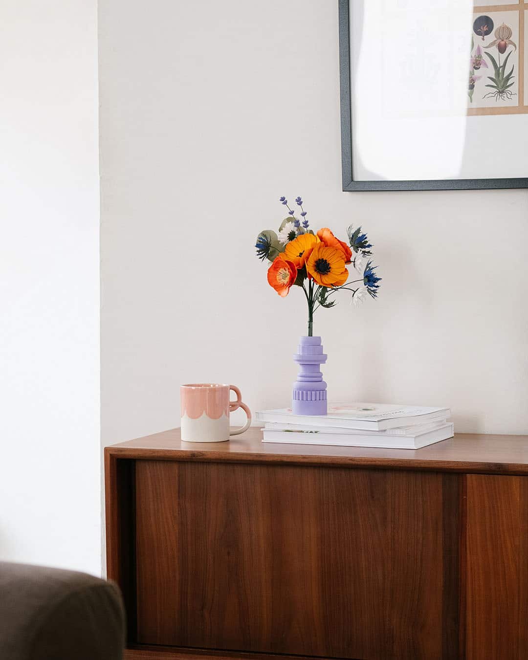Bouquet de fleurs en papier d'art Garrigue dans un vase lilas posé sur des livres, sur une commode en bois foncé, poster botanique encadré en arrière-plan, Anaïs LVDX