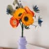 Gros plan du bouquet Garrigue avec grandes anémones jaune soleil et chardons bleus au premier plan, lavande et pâquerettes blanches visibles, vase lilas géométrique, Anaïs LVDX