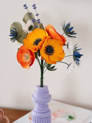 Gros plan du bouquet Garrigue avec grandes anémones jaune soleil et chardons bleus au premier plan, lavande et pâquerettes blanches visibles, vase lilas géométrique, Anaïs LVDX