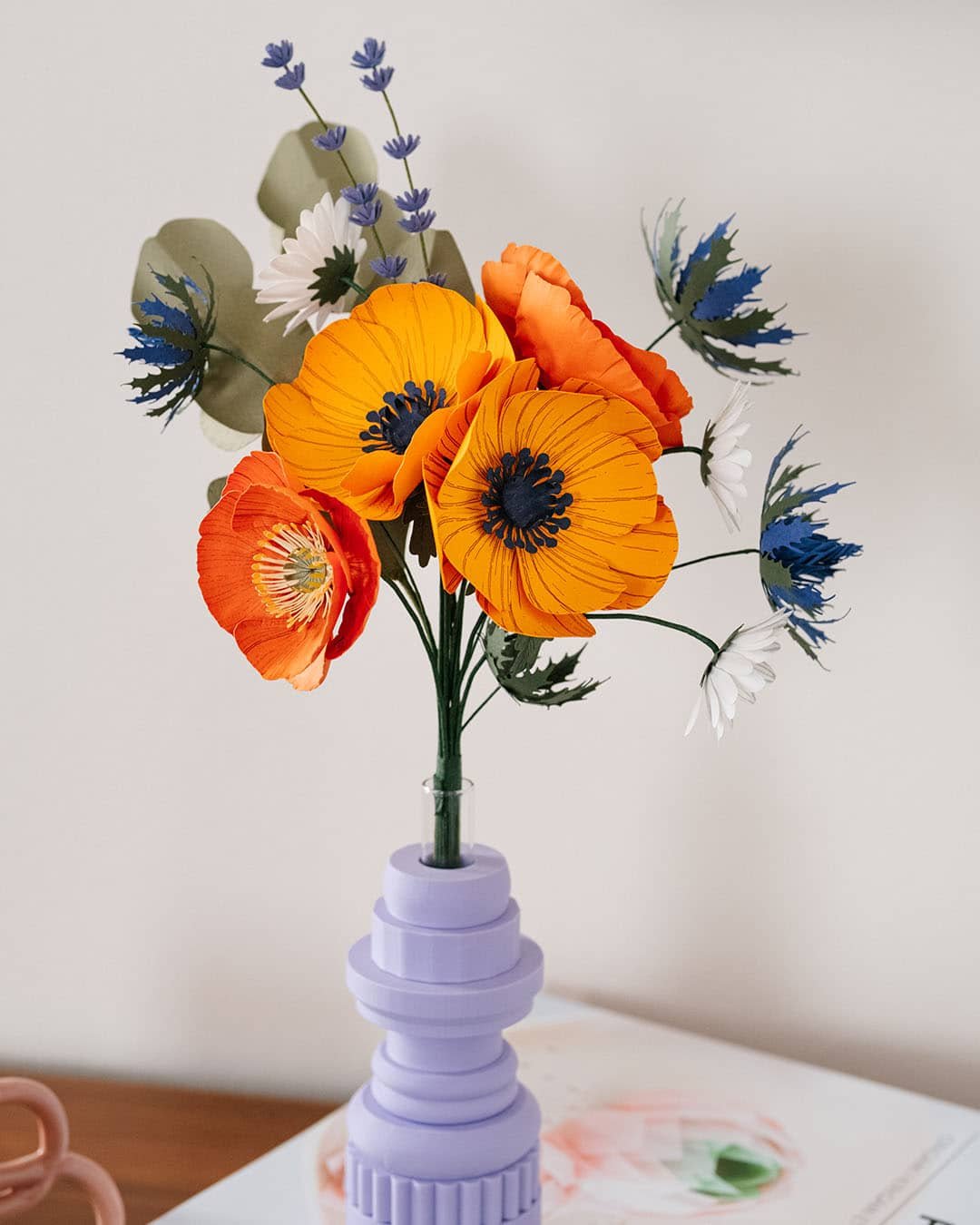 Gros plan du bouquet Garrigue avec grandes anémones jaune soleil et chardons bleus au premier plan, lavande et pâquerettes blanches visibles, vase lilas géométrique, Anaïs LVDX