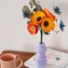 Bouquet Garrigue en papier d'art dans un vase lilas posé sur des livres, mug rose au premier plan, fond clair, anémones jaune soleil et pavots orange visibles, Anaïs LVDX