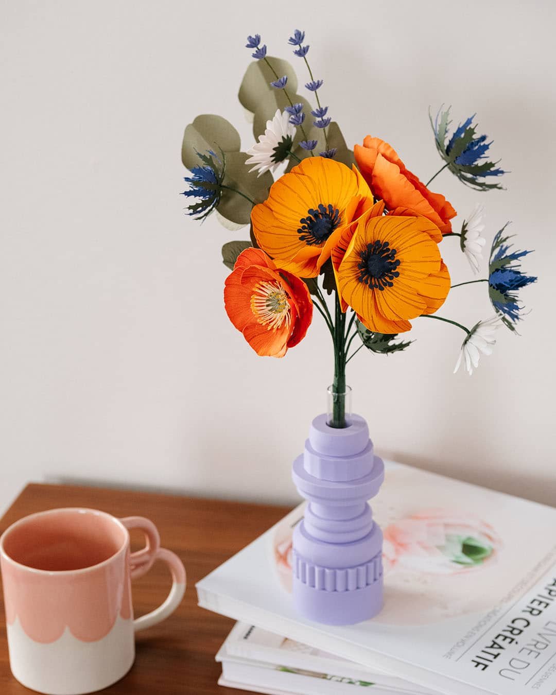 Bouquet Garrigue en papier d'art dans un vase lilas posé sur des livres, mug rose au premier plan, fond clair, anémones jaune soleil et pavots orange visibles, Anaïs LVDX