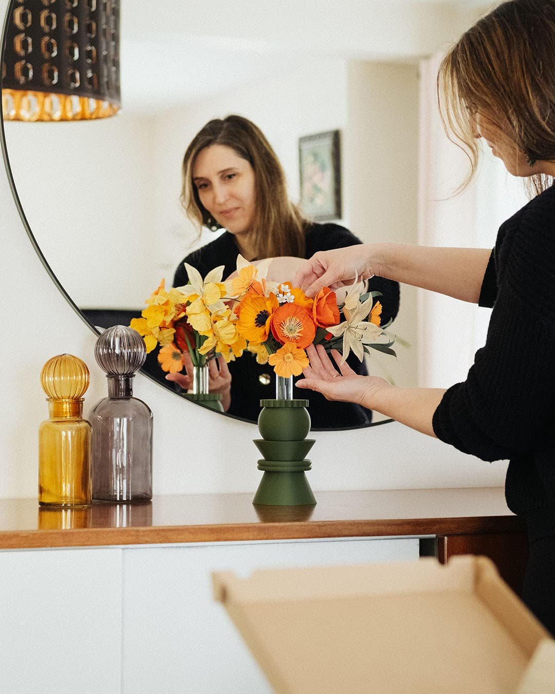 Anaïs LVDX arrange à la main son bouquet de fleurs en papier Masterpiece Orange dans un vase vert kaki, reflet dans un miroir rond