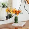 Bouquet de fleurs en papier Masterpiece Orange dans son vase vert kaki posé sur des livres sur une commode, reflet dans un grand miroir rond, décoration artisanale